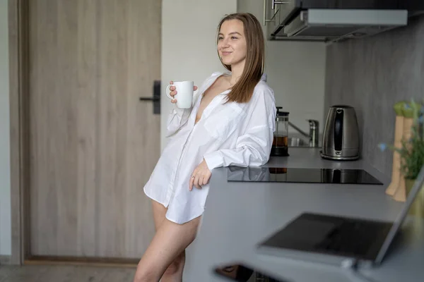 美丽的小女孩站在厨房里喝咖啡 早上例行公事穿着白衬衫 桌上的水泡式笔记本电脑和移动电话 — 图库照片