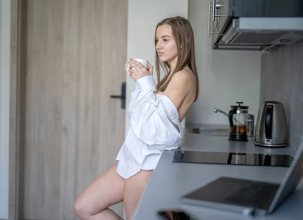 美しい若い女の子がキッチンに立ってコーヒーを飲んでいます 朝のルーチン 白いシャツを着て 机の上のブリュリーノートパソコンと携帯電話 — ストック写真