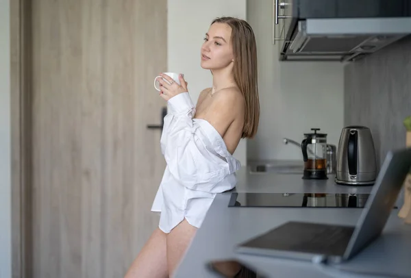 美しい若い女の子がキッチンに立ってコーヒーを飲んでいます 朝のルーチン 白いシャツを着て 机の上のブリュリーノートパソコンと携帯電話 — ストック写真