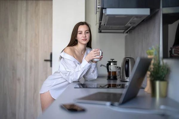 美丽的小女孩站在厨房里喝咖啡 早上例行公事穿着白衬衫 桌上的水泡式笔记本电脑和移动电话 — 图库照片