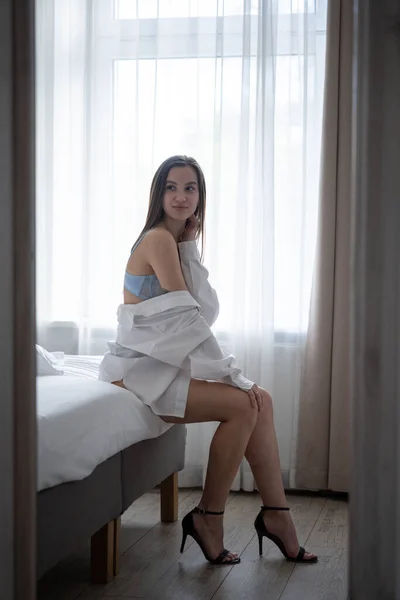 青いブラジャーと白いシャツを着た美しい若い女の子がベッドルームのベッドに座っています — ストック写真