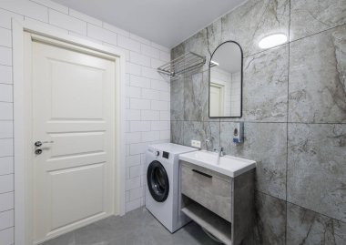 Parlak Zarif Modern Minimalist Banyo Beyaz Lavabolu Duş Odasının İç Tasarımı. Banyo aksesuarları, gri fayans duvarları, ayna, çamaşır makinesi ve musluk. LED Işık