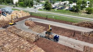 İşçi işletme yükleyicisi, modern ahşap işleme fabrikası, işleme ve imalat için yeni kesilmiş kütükleri yığına yükler. Kamyon şoförü bekliyor.