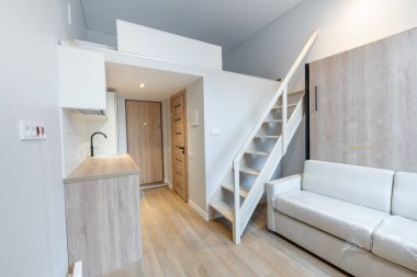 İskandinav Tarzı Minik Modern Açık Parlak Minimalizm Ev İçi. Koltuk, kanepe ve uyku alanı, mobilyalar arka planda. Koyu Kahverengi Döşeme. Beyaz Dolaplar. Yatak odasına giden merdivenler. Çatı