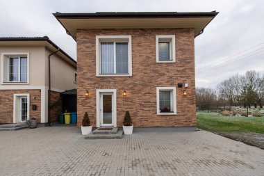 Varoş mahallesindeki iki katlı tuğladan bir evin şık cephesi iyi bakılmış garaj yolu, sıcak ışıklandırma aksanları, dingin yeşil zemin. Ev boş ve çöp kutusu dolu.