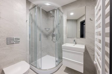 Çağdaş banyo, cam kaplı bir duş, parlak beyaz bir lavabo dolabı, gri fayanslı duvarlar, ve temiz ve minimalist bir tasarımla modern konforlar..