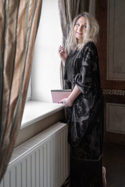 Elinde kitap tutan, koyu desenli bir pelerinle pencerenin yanında duran sessiz tonlu ve dekoratif perdeleri olan bir kadın. Huzur ve rahatlık saçan bir kadın.