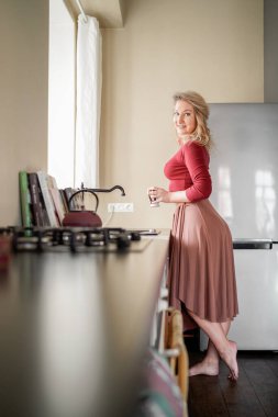 Sıcak bir mutfakta elinde kahve fincanı tutarken çıplak ayakla duran rahat bir kadın. Parlak ve rahat bir Mutfakta Sabah Kahvesinden Hoşlanan Kadın