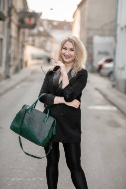 Şık giyinmiş zarif bir kadın kentsel bir arka planda modern yeşil çantayla, özgüven ve entellektüelliği yansıtıyor. Sahne kentsel moda eğilimlerini ve çağdaş yaşam tarzlarını vurguluyor