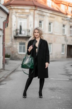 Şık giyinmiş zarif bir kadın kentsel bir arka planda modern yeşil çantayla, özgüven ve entellektüelliği yansıtıyor. Sahne kentsel moda eğilimlerini ve çağdaş yaşam tarzlarını vurguluyor