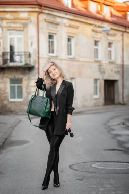 Siyah ceketli, taytlı ve topuklu ayakkabılı, yeşil çanta taşıyan şık bir kadın. Eski bir şehir bölgesinde açık havada poz vermek, özgüven yaymak ve modaya uygun estetik.