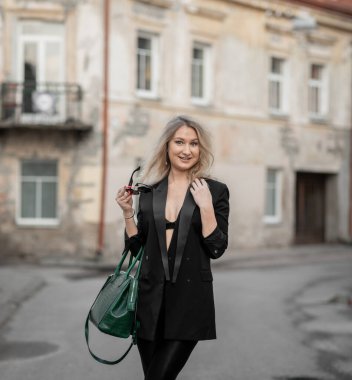 Siyah ceketli, taytlı ve topuklu ayakkabılı, yeşil çanta taşıyan şık bir kadın. Eski bir şehir bölgesinde açık havada poz vermek, özgüven yaymak ve modaya uygun estetik.