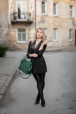 Şık bir kadın, kendine güvenen bir zarafetle bir şehir caddesinde yürüyor. Yeşil bir çanta taşıyor ve gösterişli, siyah bir kıyafet giyiyor. Şehir modasını yansıtıyor. Arka planda eski binalar