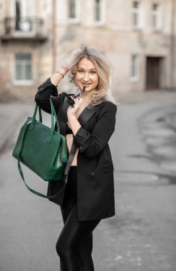 Şık bir kadın, kendine güvenen bir zarafetle bir şehir caddesinde yürüyor. Yeşil bir çanta taşıyor ve gösterişli, siyah bir kıyafet giyiyor. Şehir modasını yansıtıyor. Arka planda eski binalar