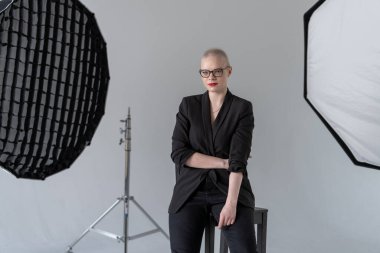 Profesyonel bir kadın, bir fotoğraf stüdyosunda, resmi iş kıyafeti giymiş, ışıklandırma ekipmanlarıyla otururken kendine güvenerek poz verir. Gülümseyen Kadın. Kanserden kurtulan. Model Glaucoma 'ya sahip