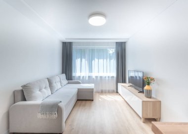 Parlak ve şık bir oturma odası, rahat bir bej kanepe, zarif bir dekor ve geniş pencerelerden yansıyan doğal aydınlatma. Kanepe, TV ve Minimalist Tasarım ile Modern Parlak Oturma Odası