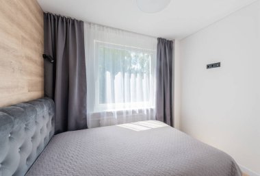 Rahat, zarif tasarlanmış yatak odası yumuşak gri bir yatak odası, ahşap elementler, gri perdeler ve modern bir pencereden gelen parlak doğal ışık. Modern Minimalist Yatak Odası İçi. Pencereler ve Perdeler