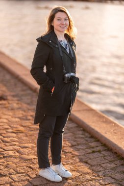 Günlük giysiler giymiş bir kadın, elinde sıcak ışıkla aydınlatılmış bir kamerayla suyun yanında duruyor. Gün batımında elinde kamerayla dışarıda duran genç bir kadın. Kopenhag, Danimarka