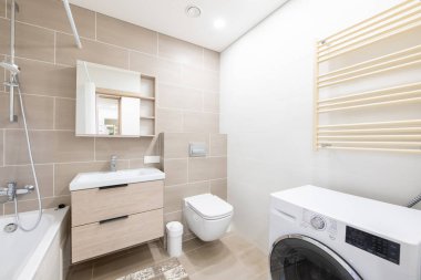 Parlak ve minimalist banyoda bej fayanslar, duvara monte edilmiş lavabo, küvet ve çamaşır makinesi. Temiz bir estetik ile günlük kullanım alanını birleştiren pratik ve işlevsel bir alan.