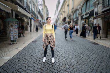 Sırt çantasıyla sıradan bir kıyafet giyen genç bir kadın Lizbon kaldırım taşı caddesinde dikiliyor. Etrafı dükkanlarla, turizm faaliyetleriyle, manzaralı mimariyle çevrili..