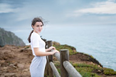 Okyanusa bakan bir çitin yanında duran bir kadının sakin görüntüsü. Kıyı manzarasının temiz havasının ve huzurunun tadını çıkarıyor. Portekiz