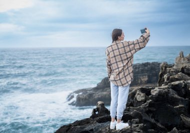 Engebeli kıyı kayalıklarında duran beyaz bir insan, arka planda geniş okyanus manzaralı bir selfie çekiyor. Portekiz