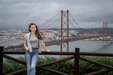 Bulutlu bir günde Lizbon 'da, ünlü bir kırmızı köprünün önünde neşeli genç bir kadın duruyor. Şehir manzarası bulanık, canlı bir zemin oluşturuyor. Sıcak bir şekilde gülümsüyor, mutluluğu ve seyahat ruhunu simgeliyor..