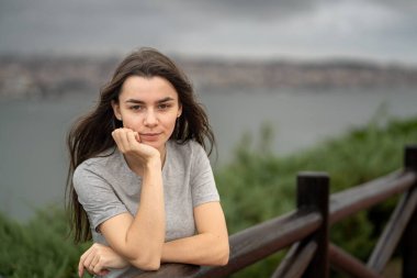 Düşünceli, genç, beyaz bir kadının portresi ahşap bir çite yaslanmış yeşil ve bulutlu gökyüzü manzaralı arka planda, huzur ve iç gözlem sunuyor. Lizbon, Portekiz