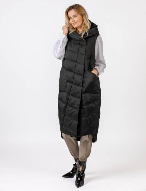 Uzun siyah bir ceketle, bej pantolon ve parlak çizmelerle donatılmış zarif bir kadın tarafsız bir arkaplanda modern bir kış kıyafeti sergiliyor..