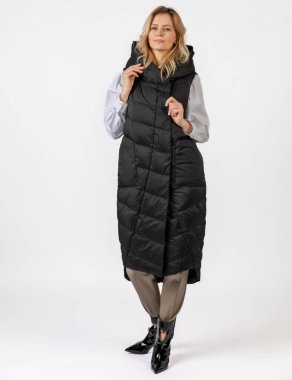 Uzun siyah bir ceketle, bej pantolon ve parlak çizmelerle donatılmış zarif bir kadın tarafsız bir arkaplanda modern bir kış kıyafeti sergiliyor..