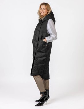 Uzun siyah bir ceketle, bej pantolon ve parlak çizmelerle donatılmış zarif bir kadın tarafsız bir arkaplanda modern bir kış kıyafeti sergiliyor..