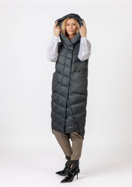 Tarafsız bir stüdyo arka planında şık bir kapüşonlu uzun yorgan giymiş, şık giyinmiş, şık giyinmiş ve gülümseyen bir kadın..