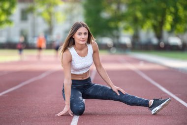 Genç beyaz bir kadın gündüz vakti koşu pistinde esneme egzersizleri yapıyor. Atletik giyinmiş, odaklanmayla rahatlamayı birleştiriyor.