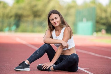 Genç beyaz bir kadın atletik bir pistte oturur, dışarıda egzersiz yaptıktan sonra rahatlamış görünür..