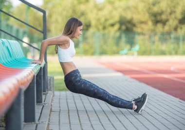 Kafkas atletik genç bir kadın parktaki bankta egzersiz yapıyor, kol gücü için doğal ışık altında triseps yapıyor..
