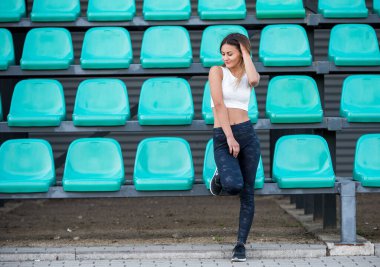 Kendine güvenen, atletik giysili beyaz bir kadın açık hava mekanında yeşil plastik koltuklara yaslanıyor. Bu resim fitness, rahatlama ve açık hava anlarının temalarını iletir.