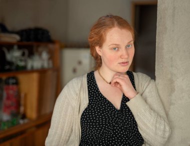 Kapalı alanda uzun kızıl kızıl saçlı, güzel ve barışçıl ifadesini vurgulayan genç bir kadını gösteren sakin bir portre..