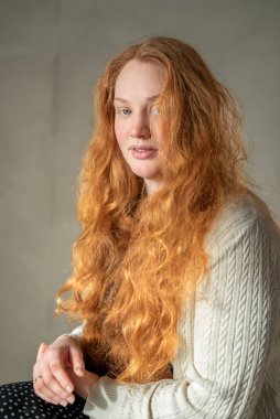 Kızıl saçlı, genç, beyaz saçlı, kıvırcık saçlı bir kadının portresi yumuşak doğal ışığın altında duruyor, kameraya bakarken özgüven ve zarafetini gösteriyor..