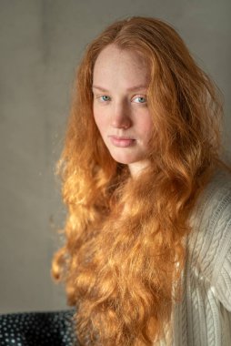 Kızıl saçlı, genç, beyaz saçlı, kıvırcık saçlı bir kadının portresi yumuşak doğal ışığın altında duruyor, kameraya bakarken özgüven ve zarafetini gösteriyor..