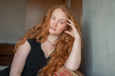 Kızıl saçlı, kapalı mekanlı, rahat bir şekilde oturan, doğal güzellik, cazibe ve rahatlama hissi saçan sakin ve düşünceli bir kadın portresi..