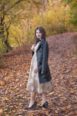 Zarif giyimli bir kadın güz ortamında yapraklarla kaplı bir yolda güven içinde durur. Etrafını çevreleyen canlı yeşillik ve şık tavırları ılık bir mevsimsel portre yaratıyor dinginlik ve cazibe..