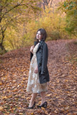 Zarif giyimli bir kadın güz ortamında yapraklarla kaplı bir yolda güven içinde durur. Etrafını çevreleyen canlı yeşillik ve şık tavırları ılık bir mevsimsel portre yaratıyor dinginlik ve cazibe..