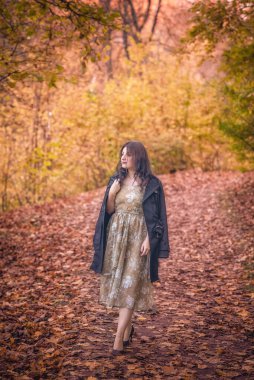 Beyaz, beyaz, şık bir elbise giymiş, paltolu bir kadın turuncu ve sarı yapraklarla çevrili, canlı bir sonbahar yolunda yürüyor..