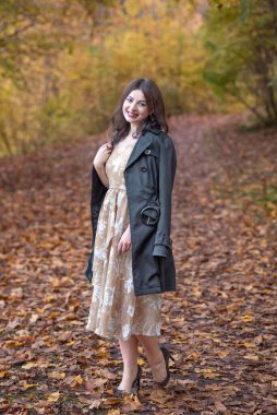 Zarif giyimli bir kadın güz ortamında yapraklarla kaplı bir yolda güven içinde durur. Etrafını çevreleyen canlı yeşillik ve şık tavırları ılık bir mevsimsel portre yaratıyor dinginlik ve cazibe..
