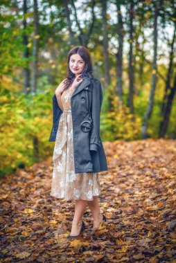 Zarif giyimli bir kadın güz ortamında yapraklarla kaplı bir yolda güven içinde durur. Etrafını çevreleyen canlı yeşillik ve şık tavırları ılık bir mevsimsel portre yaratıyor dinginlik ve cazibe..