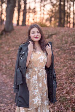 Çiçekli bir elbise ve ceket giyen alımlı bir kadın gün batımında manzaralı bir ortamda duruyor, sonbahar yapraklarıyla çevrili, doğal bir ortamda zarafet ve sükuneti ifade ediyor..