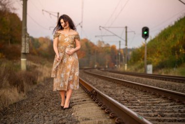 Çiçekli bir gece elbisesi giymiş zarif bir kadın gün batımında boş tren raylarında duruyor, sakin bir sonbahar manzarasında romantizm ve yalnızlık hissi yakalıyor..