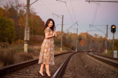 Çiçek desenli bir elbise giymiş bir kadın, sonbaharda demiryolu raylarında zarif pozlar verir. Etrafı renkli ağaçlar ve yumuşak bir atmosfer parıltısıyla çevrilidir..