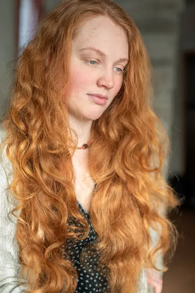 Kapalı alanda uzun kızıl kızıl saçlı, güzel ve barışçıl ifadesini vurgulayan genç bir kadını gösteren sakin bir portre..