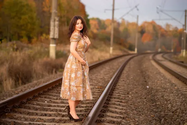 Çiçek desenli bir elbise giymiş bir kadın, sonbaharda demiryolu raylarında zarif pozlar verir. Etrafı renkli ağaçlar ve yumuşak bir atmosfer parıltısıyla çevrilidir..